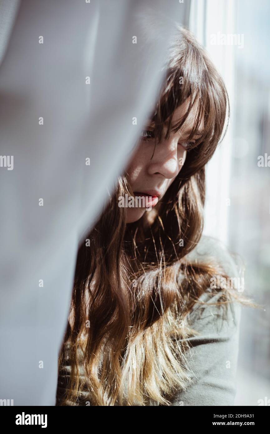 Close-up of young woman hiding behind window curtain Stock Photo