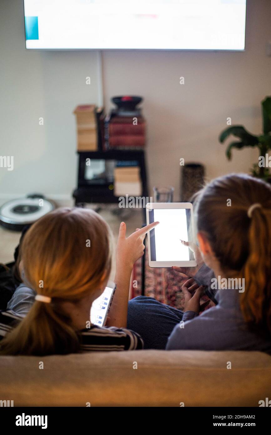 Rear view of sisters using portable information device while sitting in living room at home Stock Photo