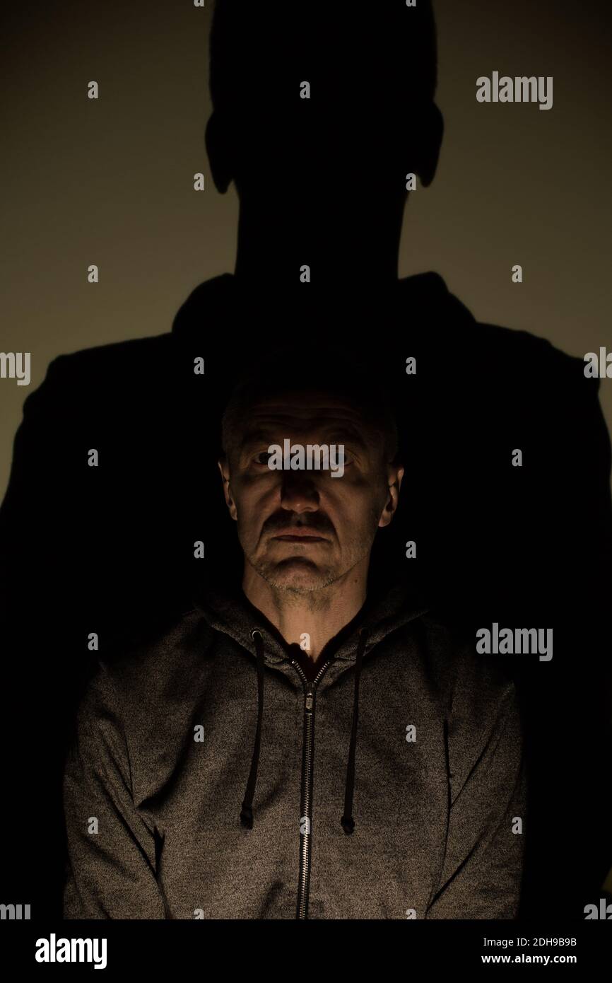 Mid adult man wearing hoodie with big shadow behind looking at the camera. Mental disorder conceptual photography. Stock Photo