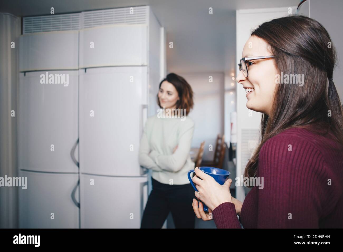 Side view of happy woman with coffee cup standing against female friend in dorm Stock Photo