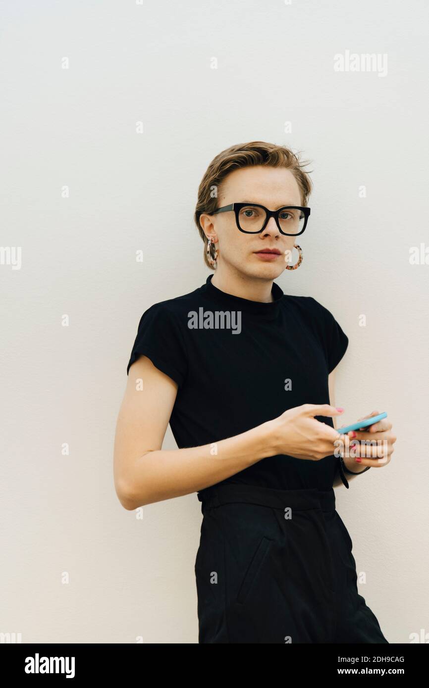 Confident androgynous entrepreneur standing with smart phone against while wall in board room Stock Photo