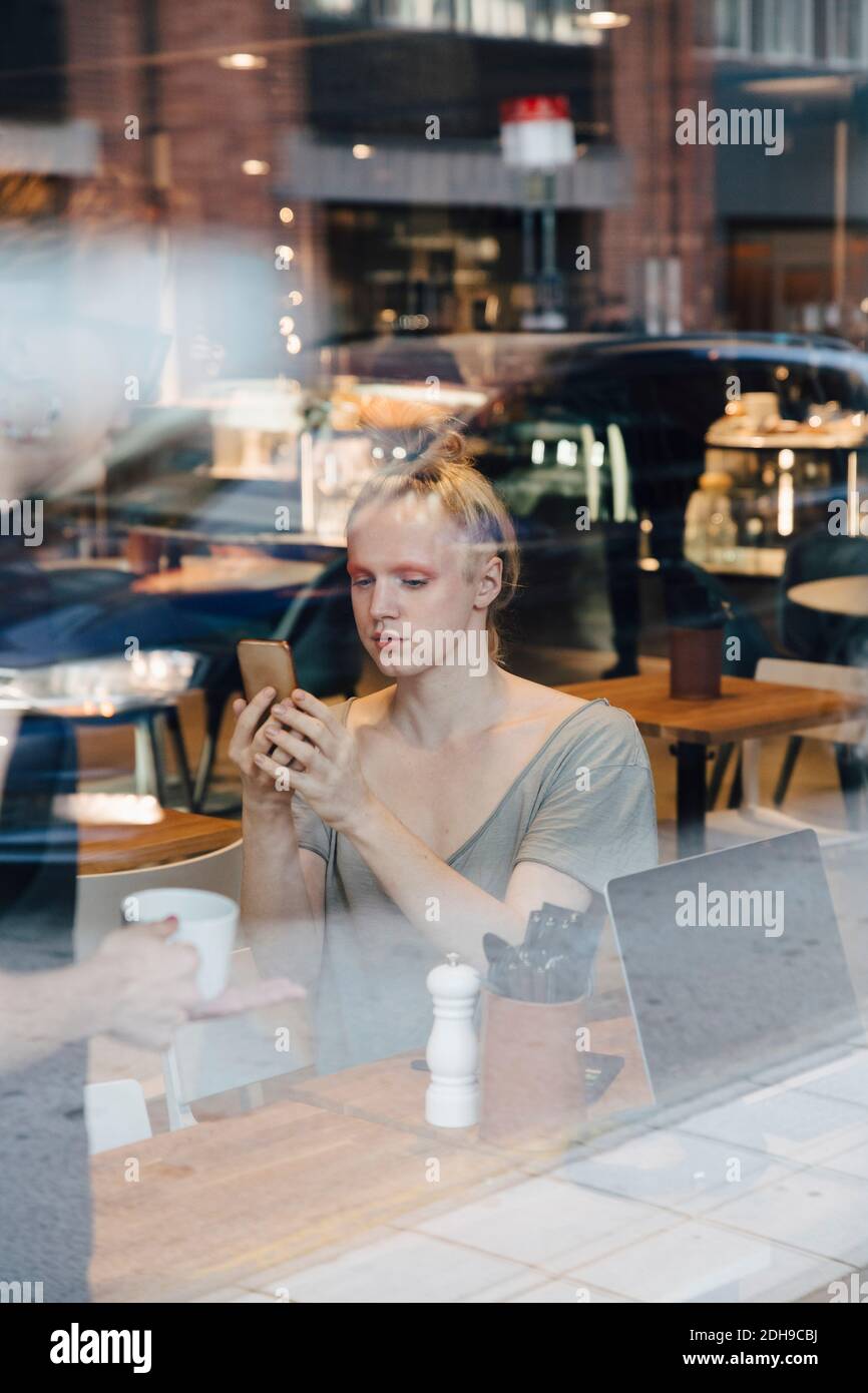Young androgynous customer using smart phone while sitting in cafe seen from window Stock Photo