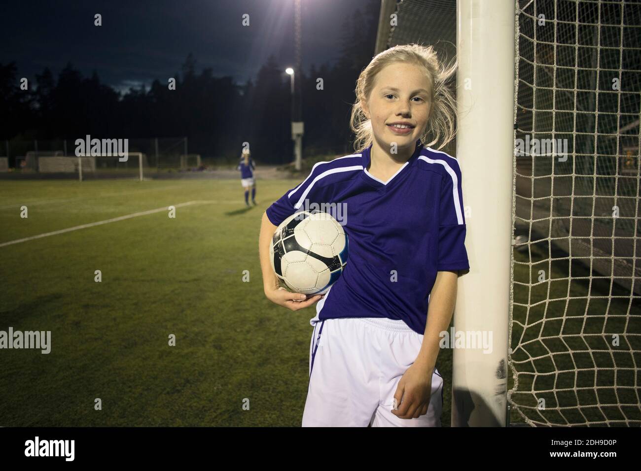 Portrait of happy girl holding soccer ball standing by goal post on field Stock Photo