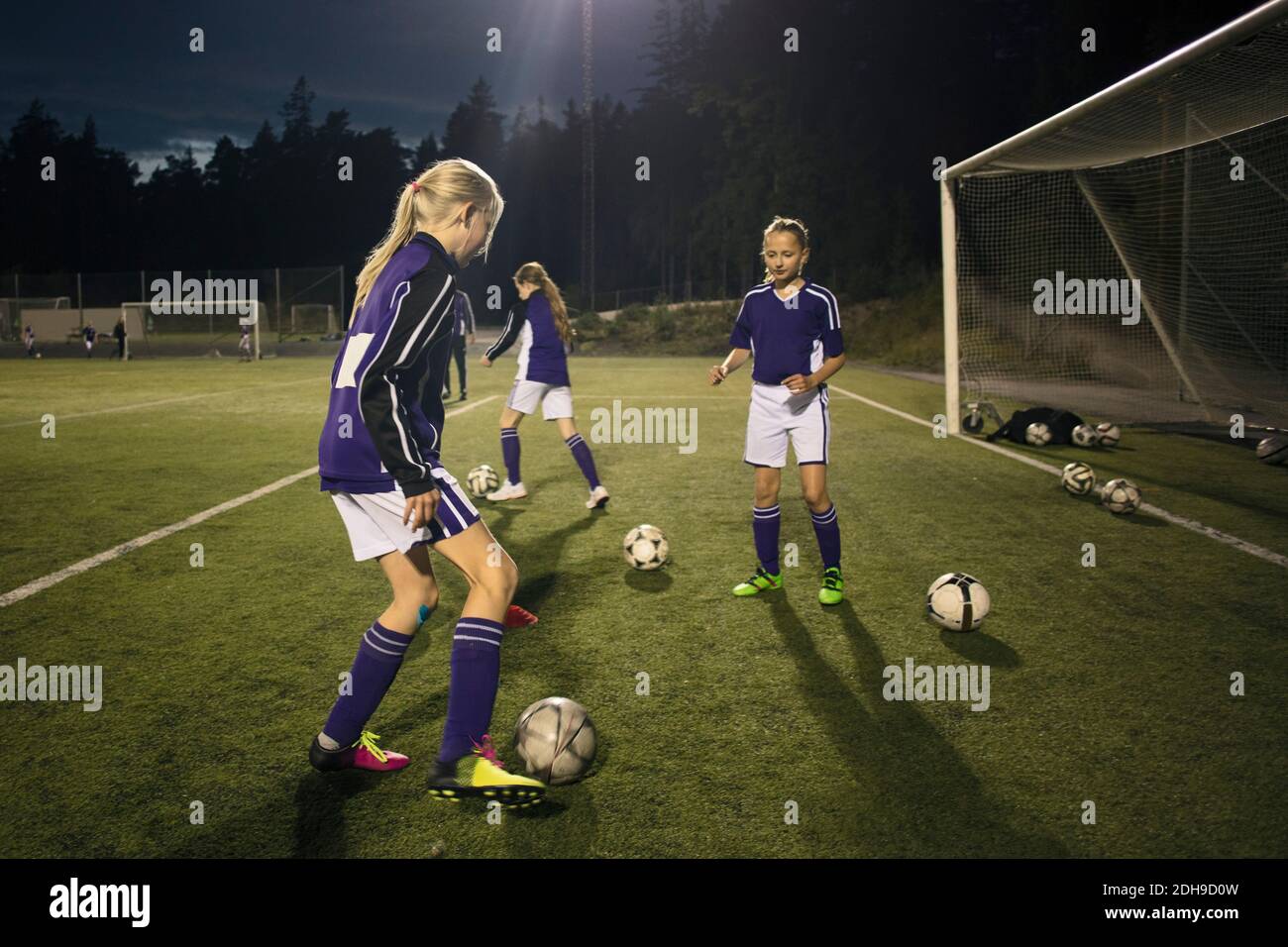 Girls practicing with soccer balls by goal post on field at night Stock Photo