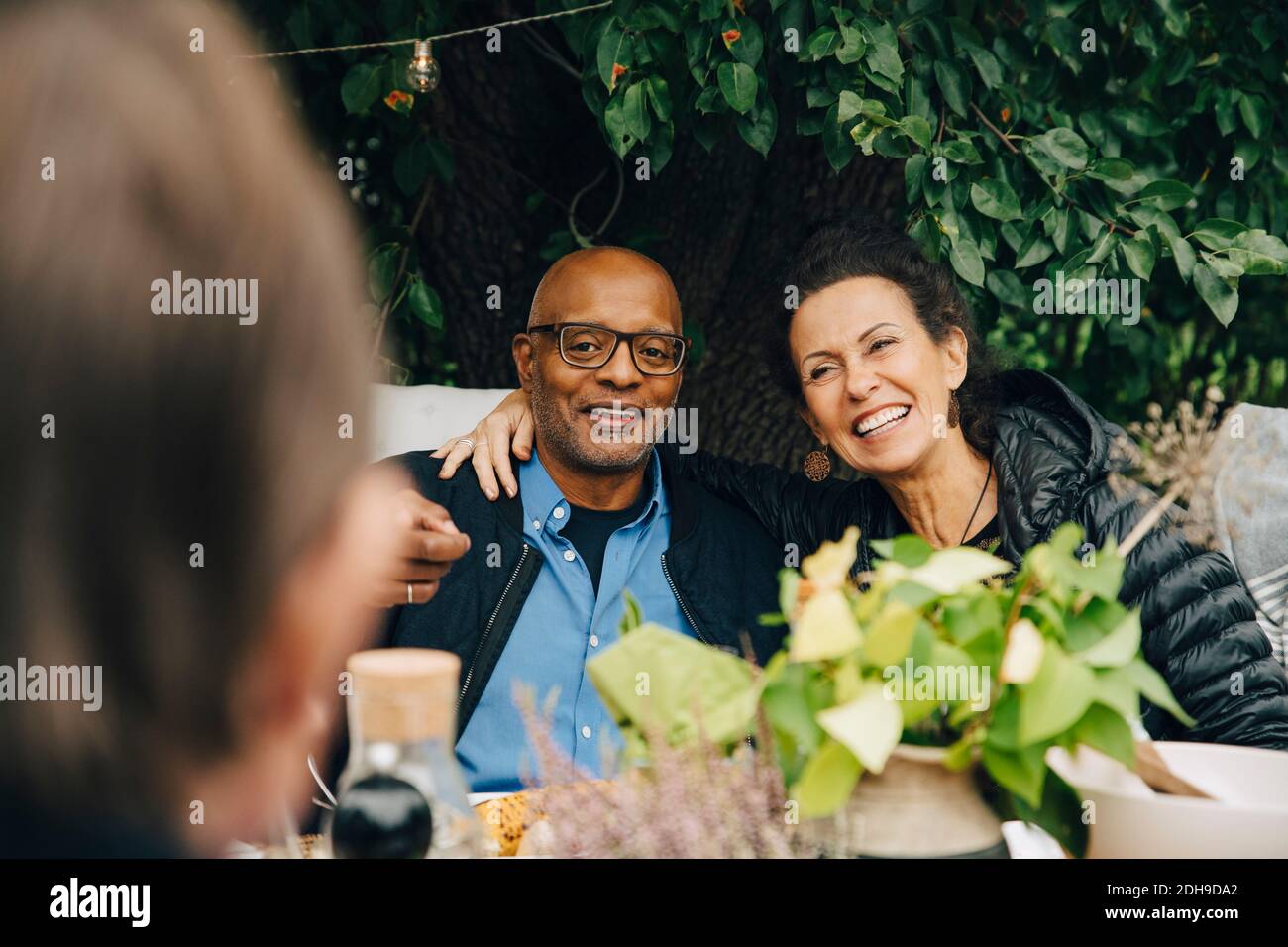Cheerful senior woman sitting with arm around man while enjoying dinner party at back yard Stock Photo