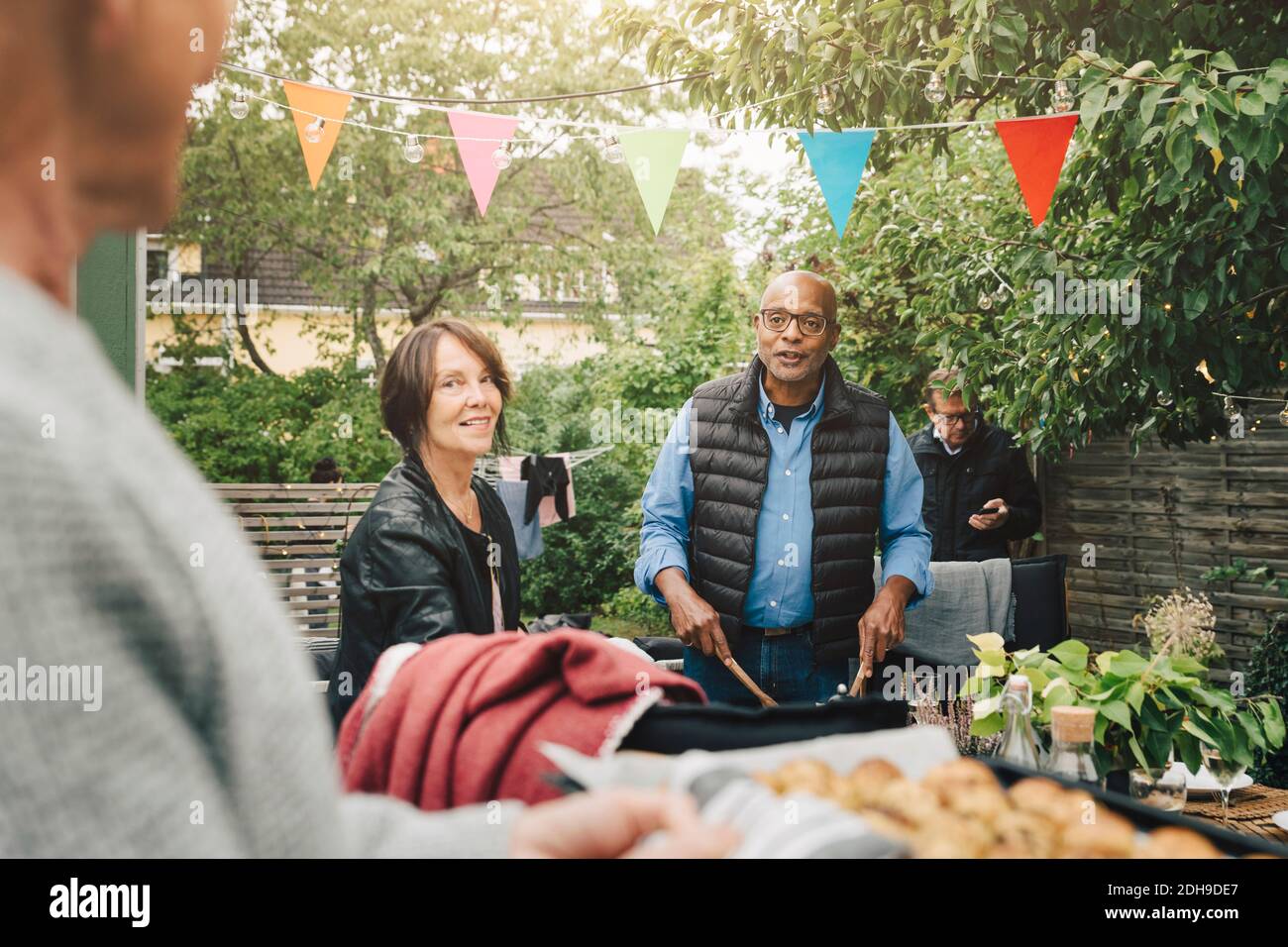 Senior friends looking at man bringing baked potatoes during dinner party at back yard Stock Photo