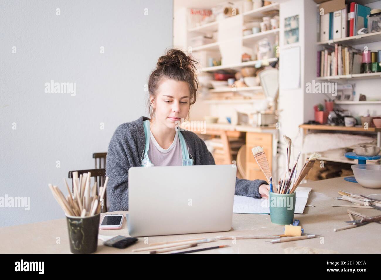 Young female potter using laptop while sitting at table in workshop Stock Photo