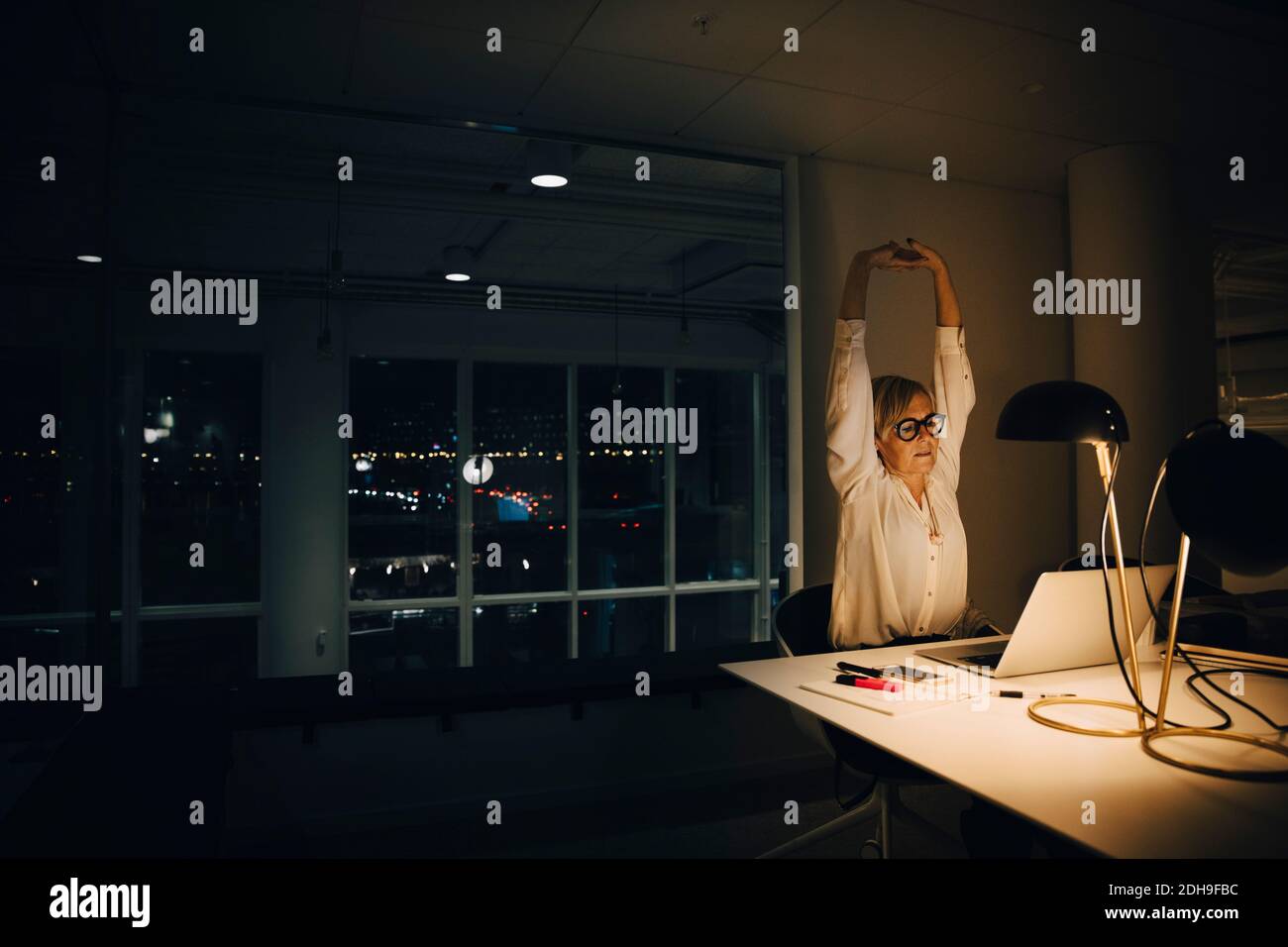 Exhausted businesswoman stretching with arms raised while working late in illuminated coworking office Stock Photo
