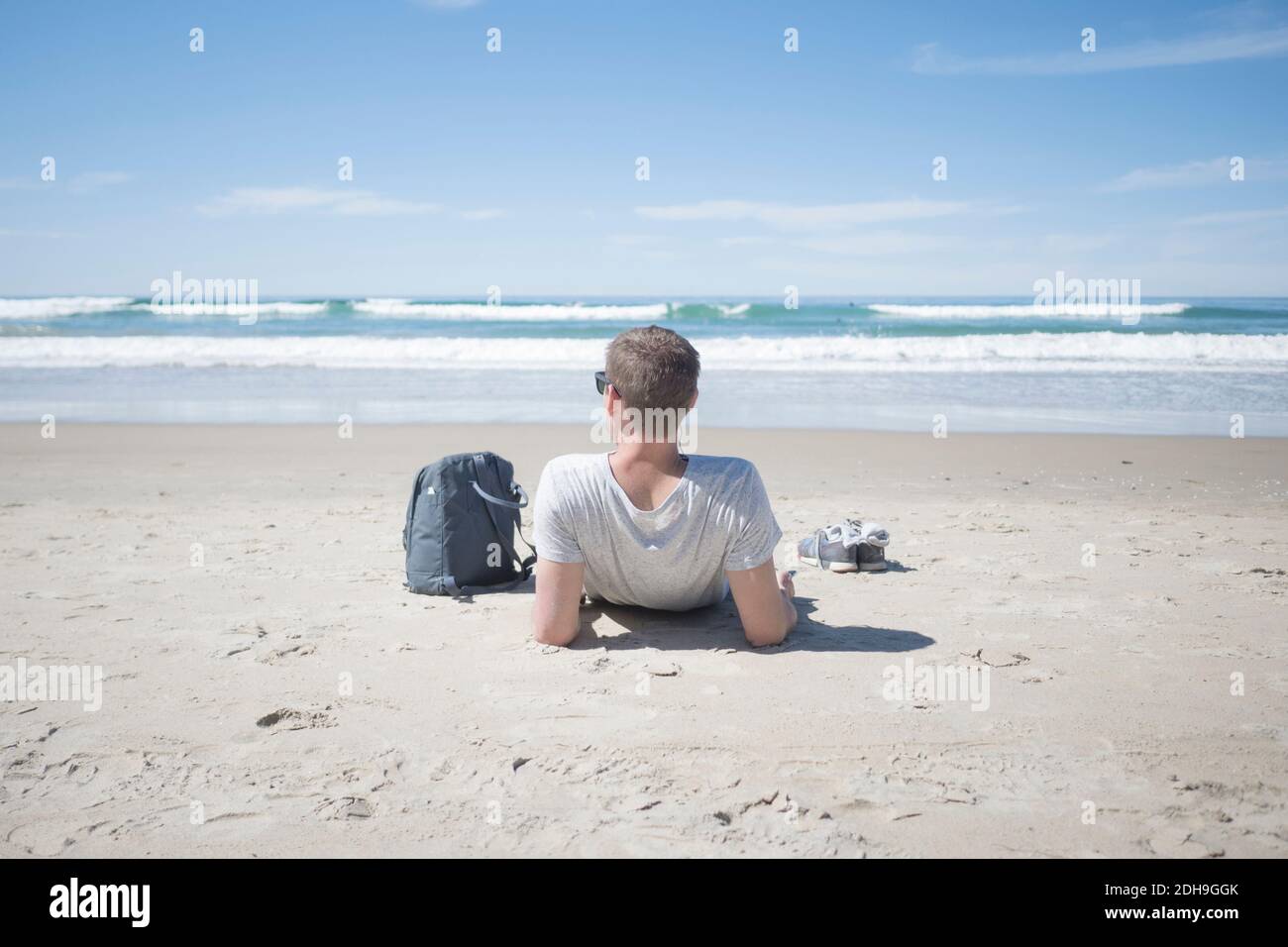 Rear view of man relaxing by backpack at beach on sunny day Stock Photo