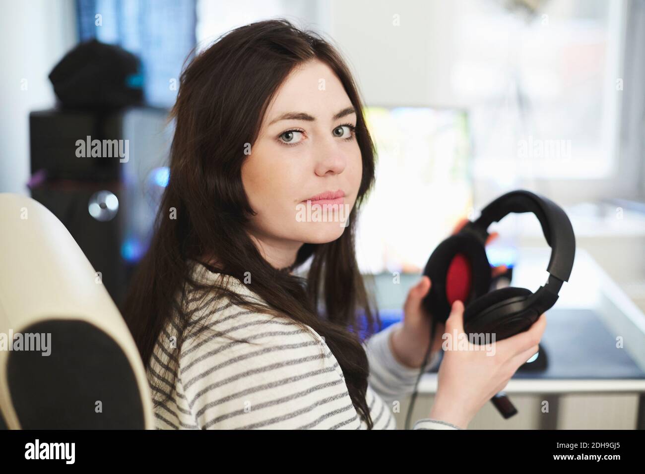 Portrait of young woman holding headphones sitting on chair at home ...