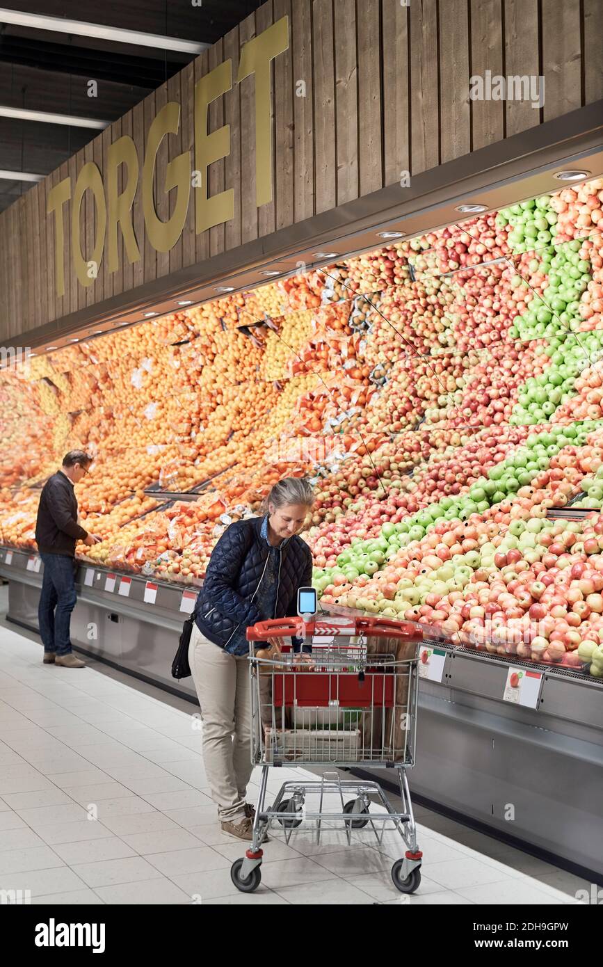 Woman standing by shopping cart while man buying apples from rack at supermarket Stock Photo