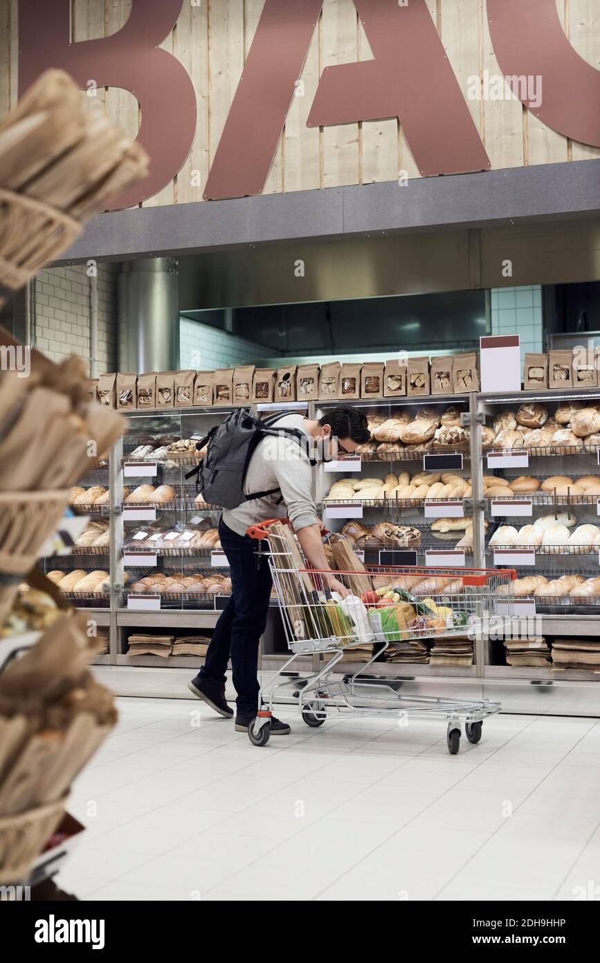Man keeping paper bag in shopping cart while standing by bread rack at supermarket Stock Photo