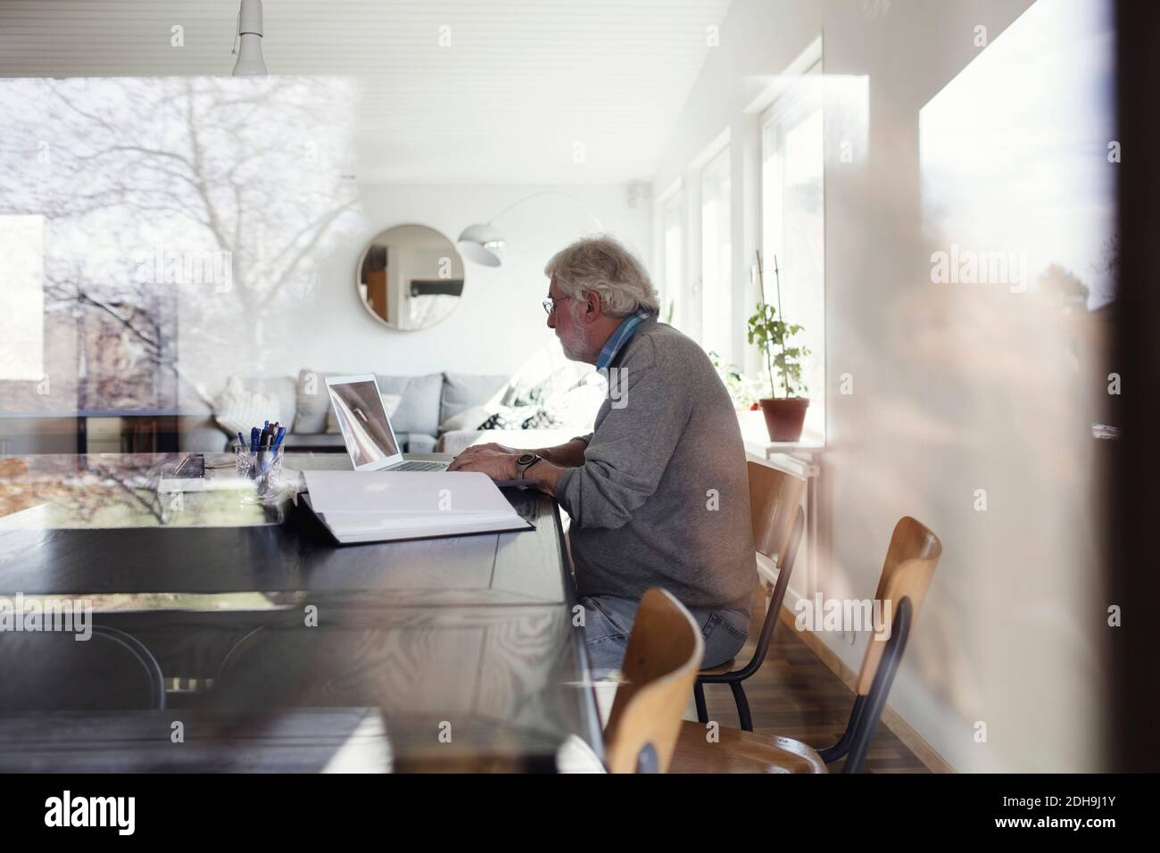 Senior man using laptop while sitting at table seen through window Stock Photo