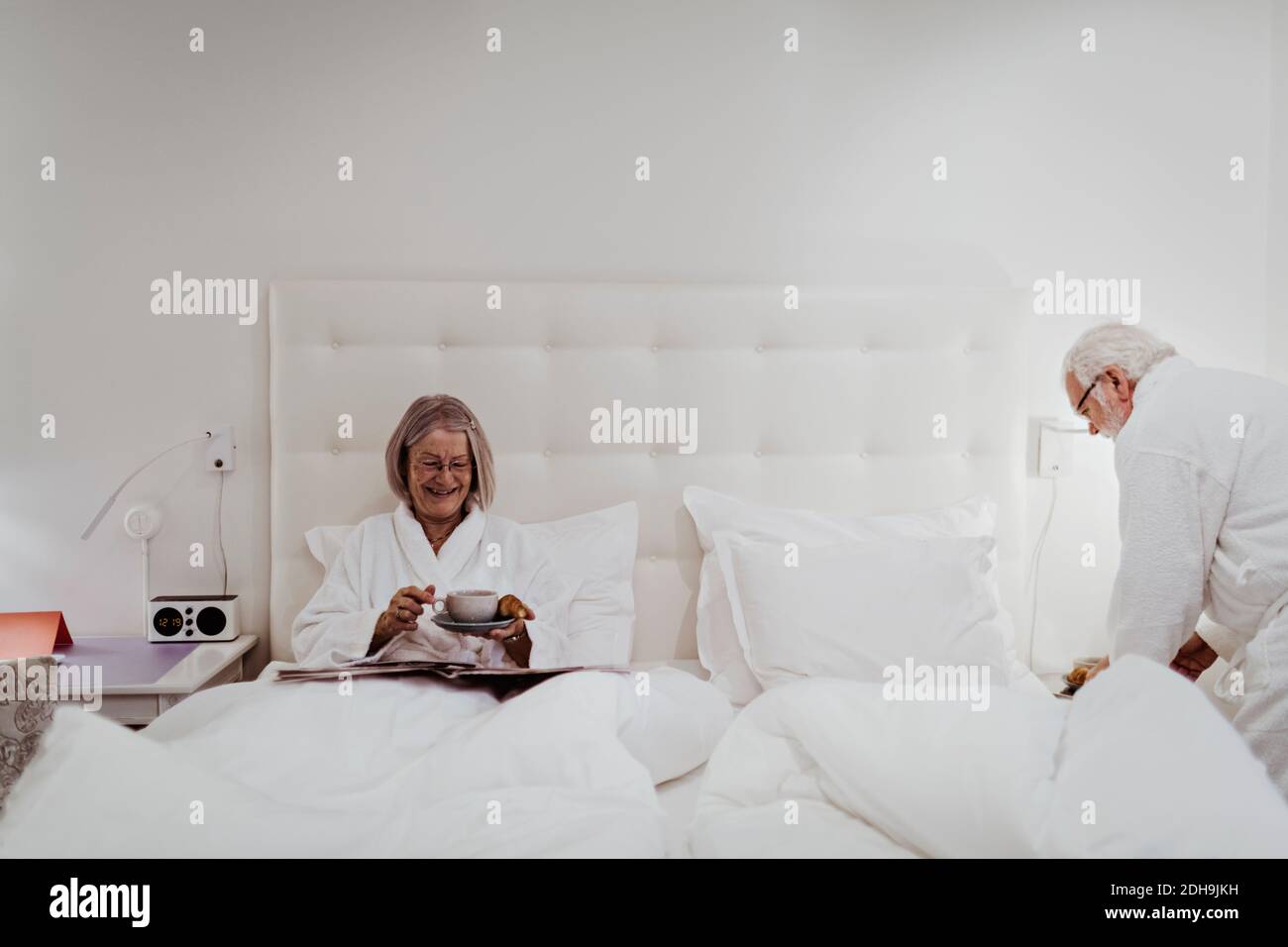Happy senior woman having coffee while man standing by bed in hotel room Stock Photo