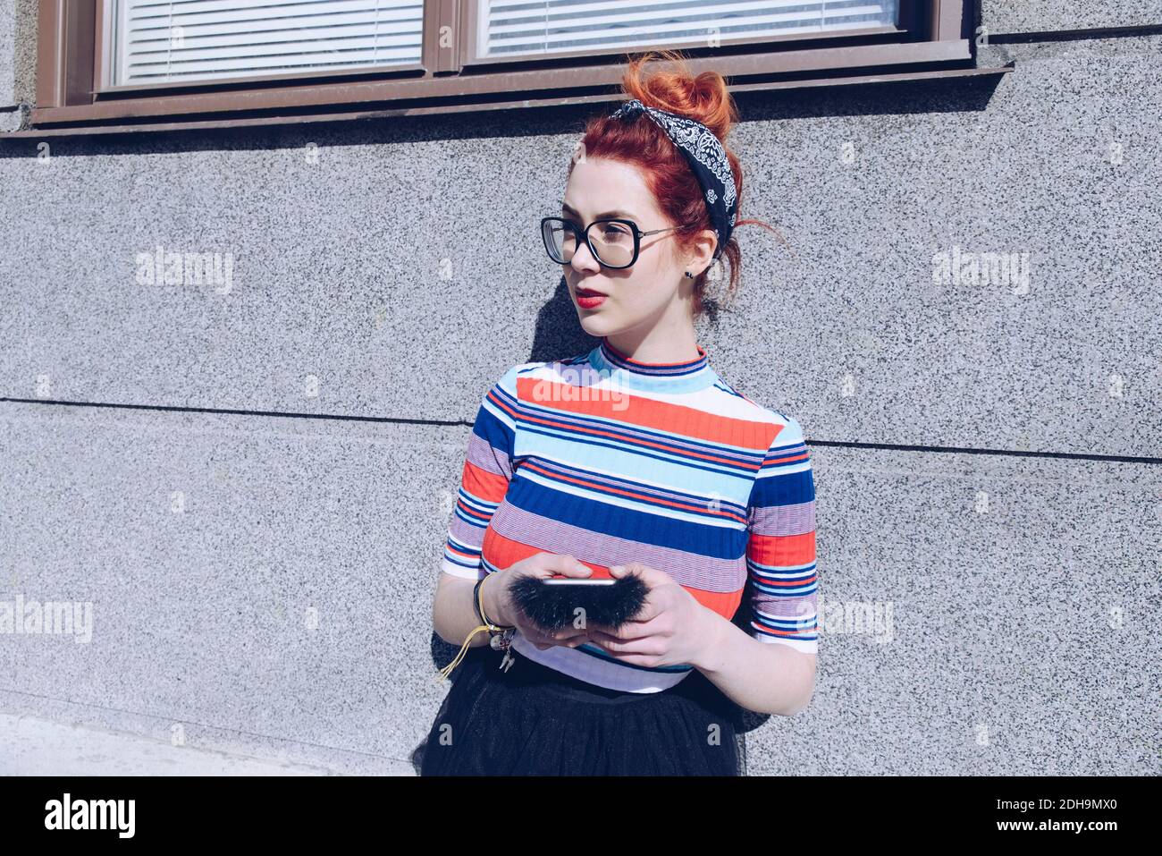 Young female hipster looking away while using mobile phone against wall Stock Photo