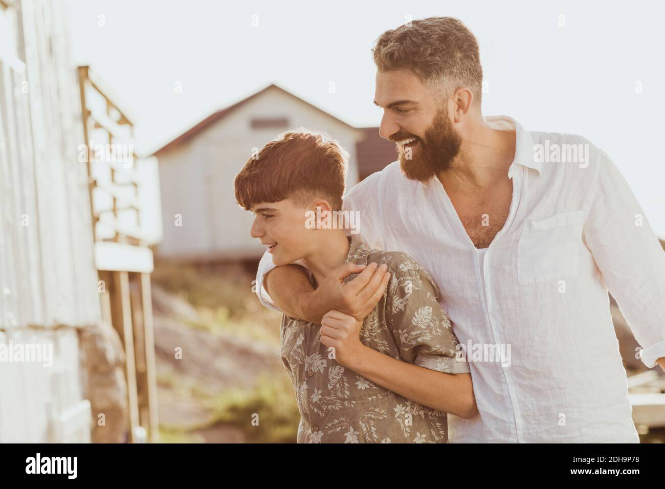 Smiling father embracing teenage son while standing against sky during sunny day Stock Photo
