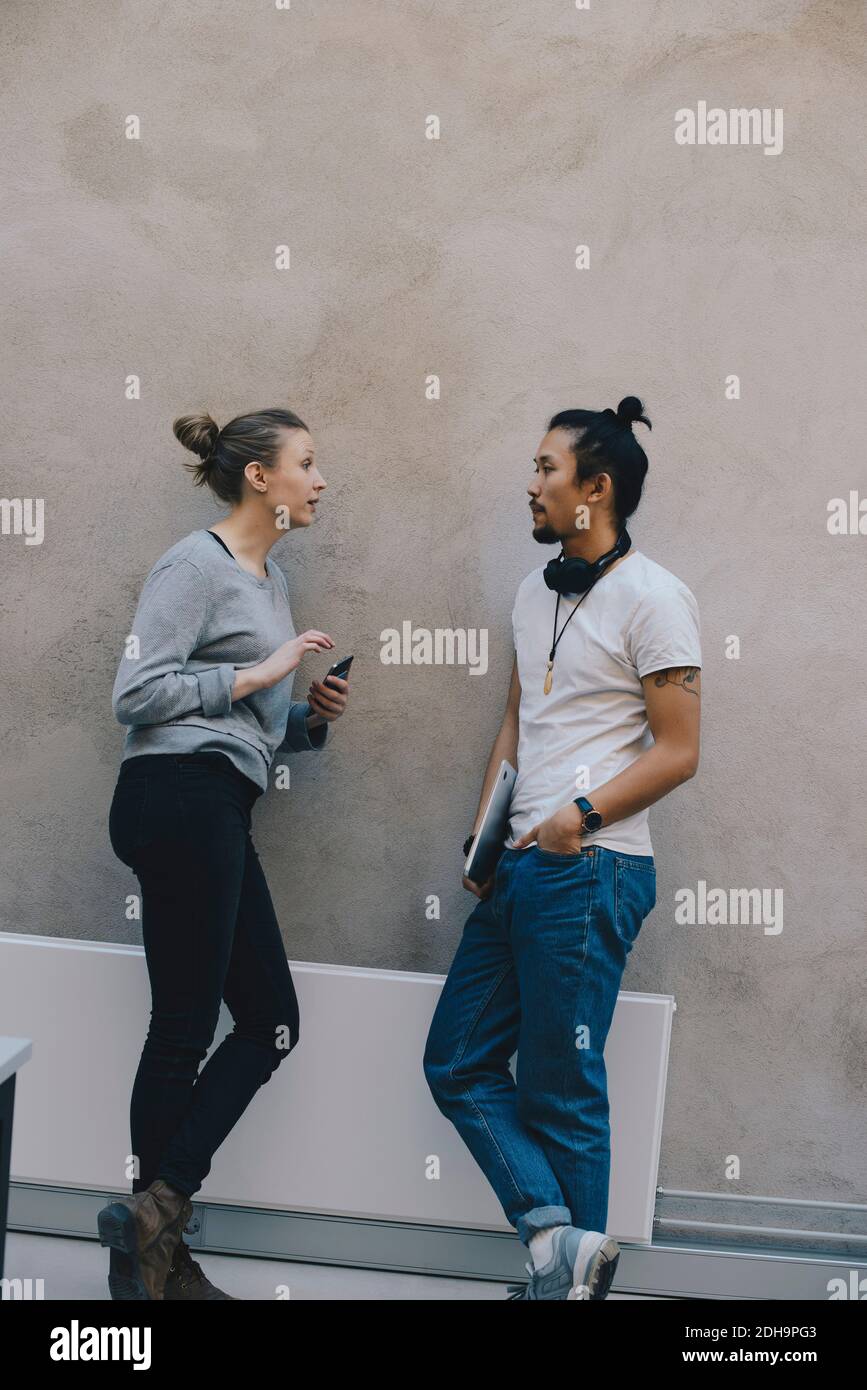 Male and female computer programmers discussing while leaning on wall in office Stock Photo