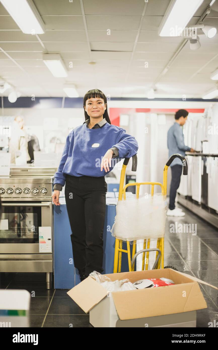 Portrait of smiling saleswoman standing by luggage cart in electronics store Stock Photo