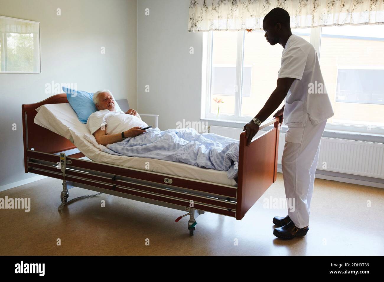 Male nurse adjusting bed while senior man watching TV in hospital ward Stock Photo