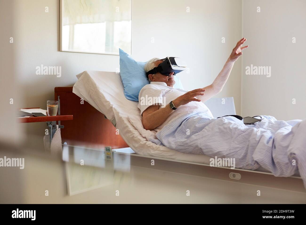 Senior man gesturing while using VR glasses on bed in hospital ward Stock Photo