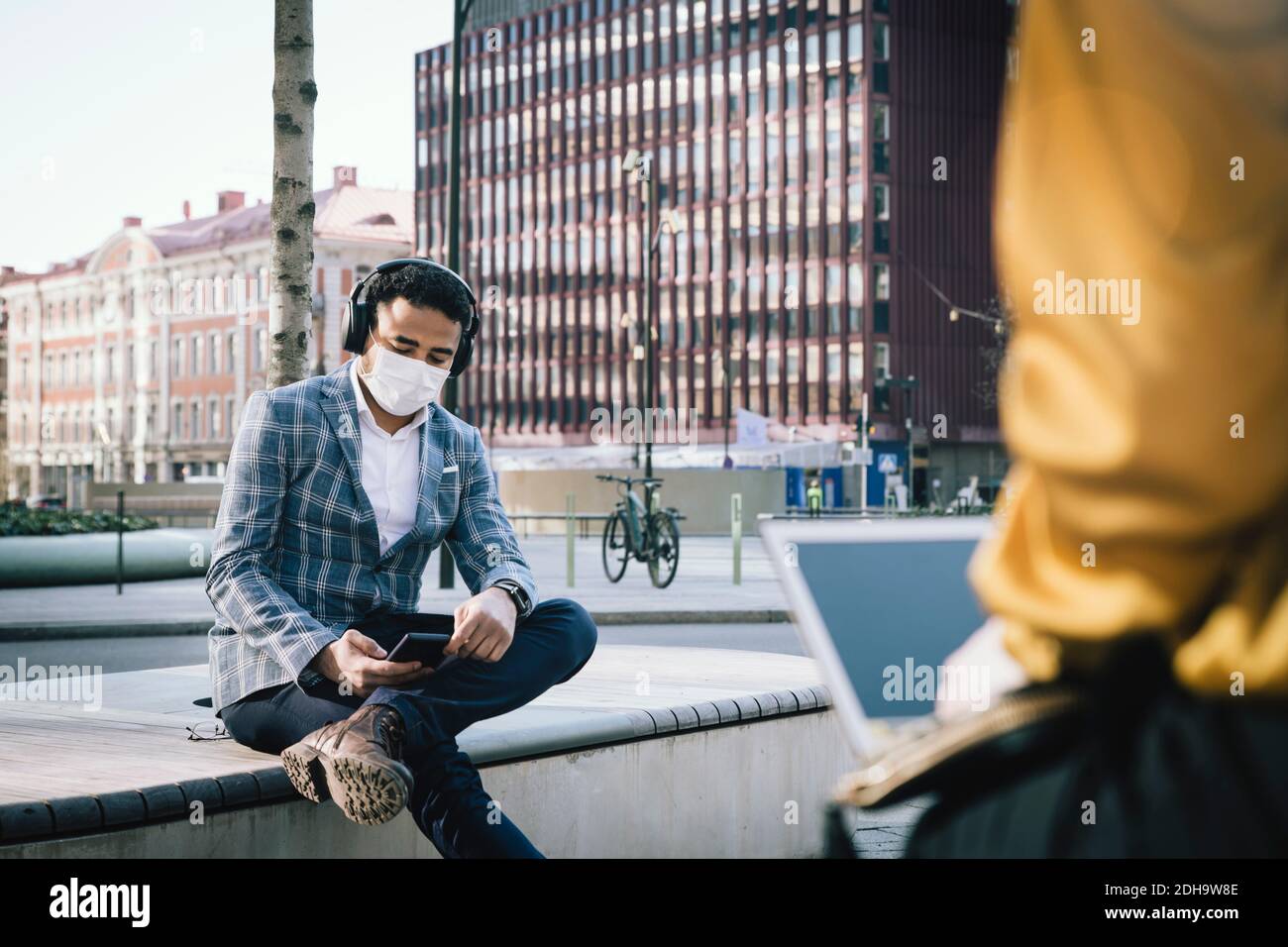 Businessman wearing face mask sitting outside using smartphone Stock Photo