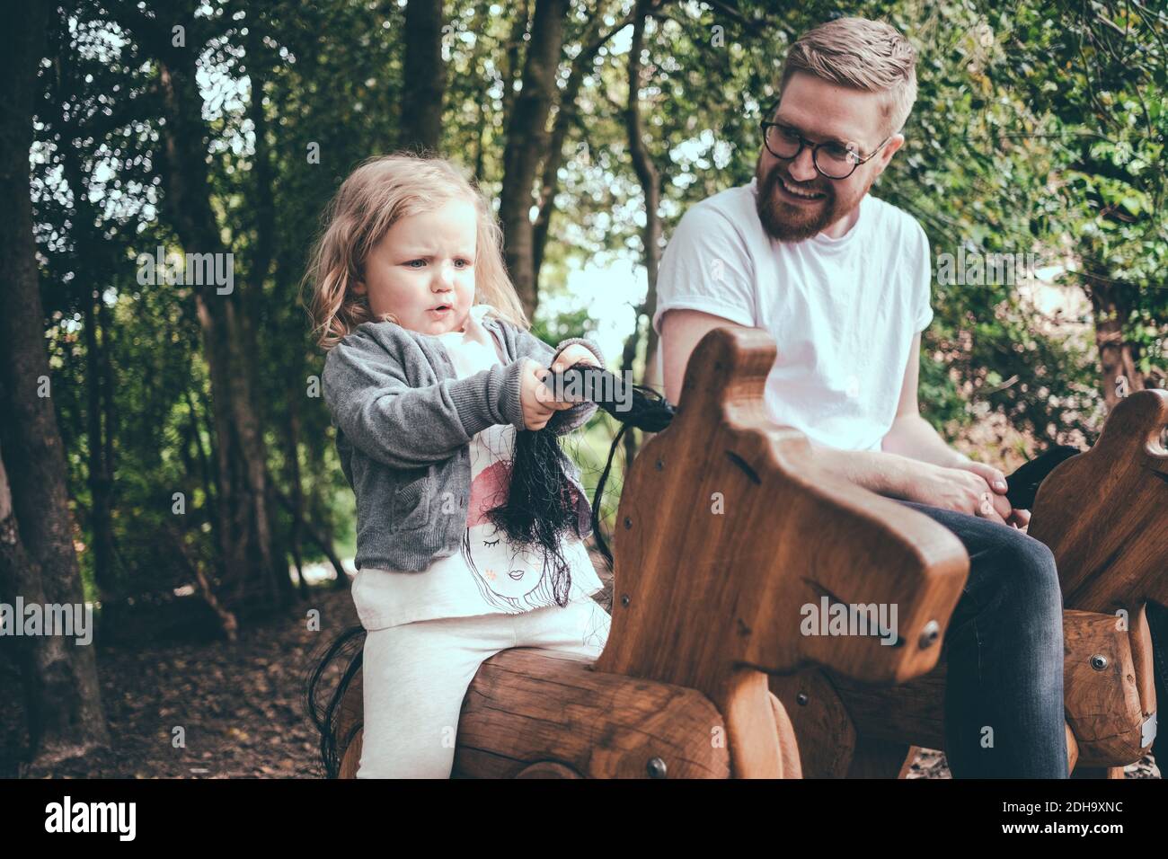 Happy father looking at daughter riding wooden horse in park Stock Photo