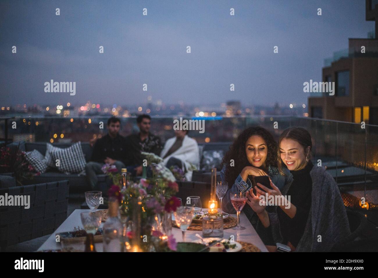 Smiling woman showing smart phone to female while friends sitting in background during social gathering on building terr Stock Photo