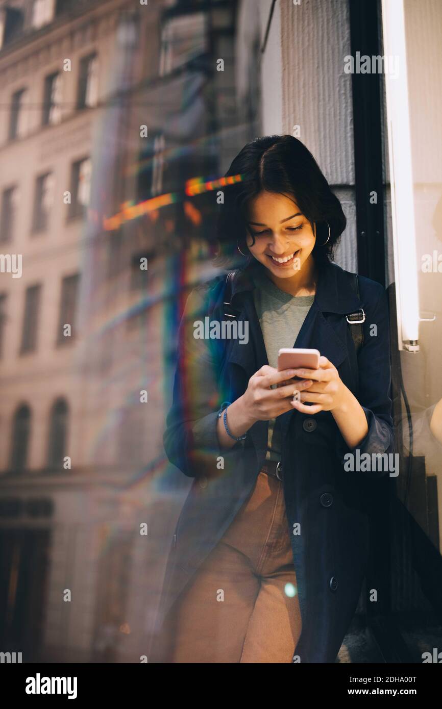 Smiling young woman texting through phone while standing by glass window Stock Photo