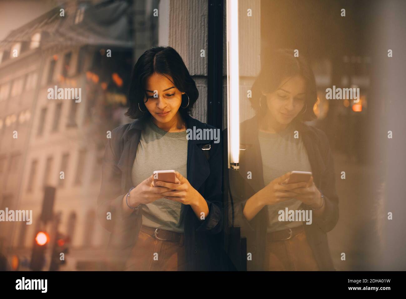 Texting Through Phone Young woman texting through phone while standing by glass window Stock ...