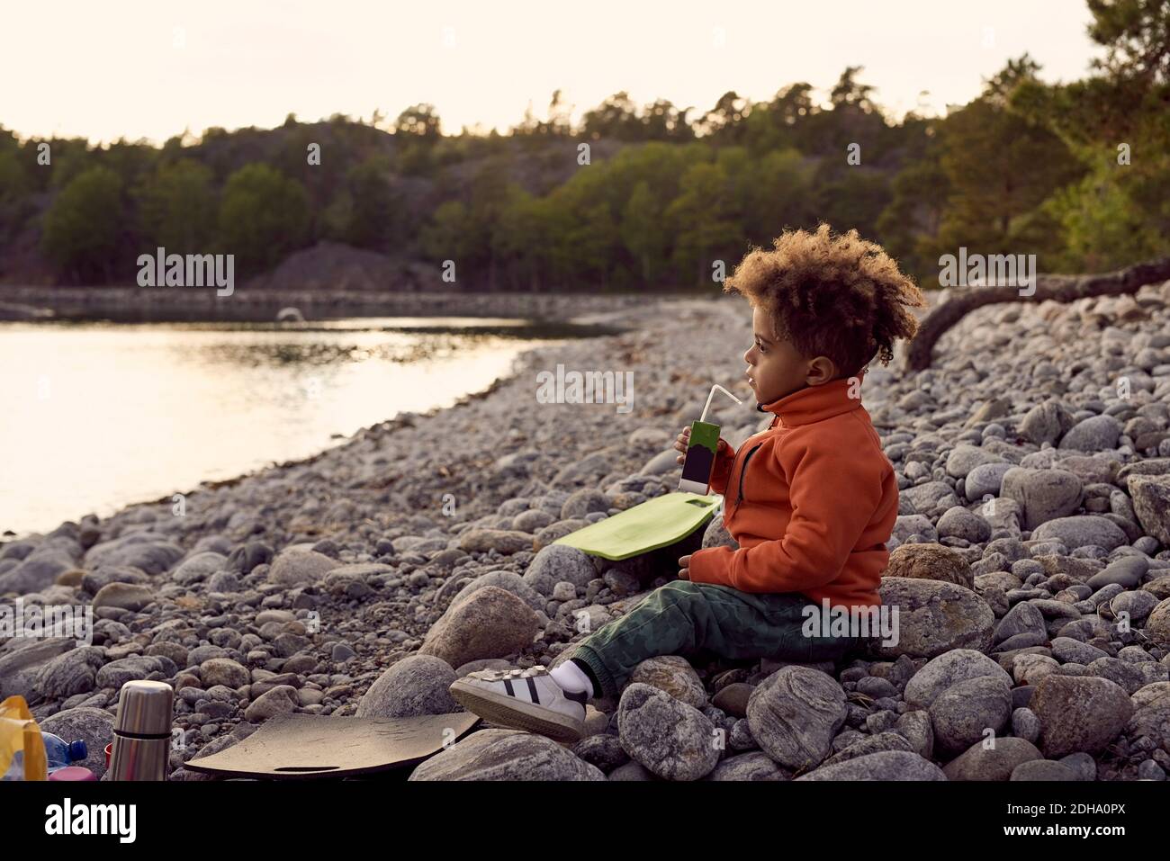 Side view of boy drinking while sitting on rocks at beach during sunset Stock Photo