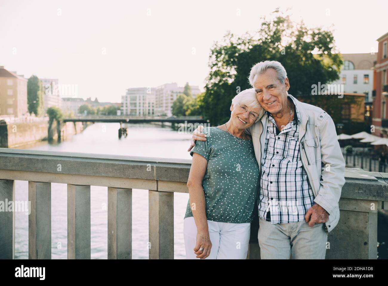 Portrait of smiling senior couple standing arm around against railing in city Stock Photo