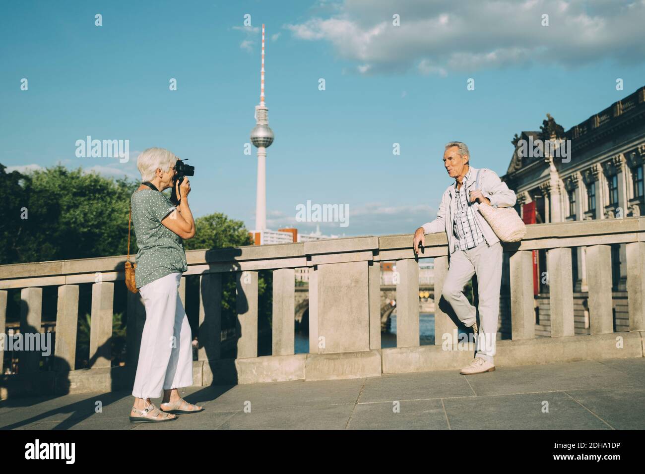 Full length of woman taking photograph of senior man while standing on bridge against tower in city Stock Photo