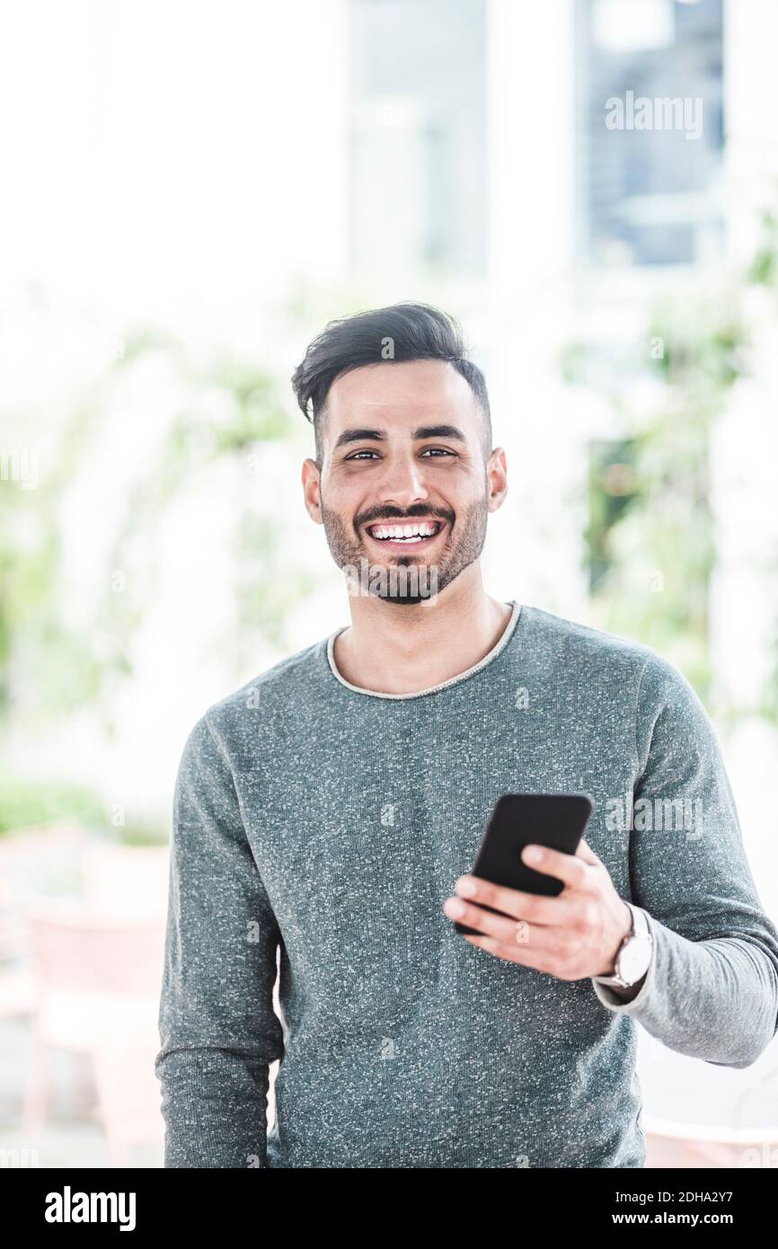 Portrait of smiling businessman holding mobile phone while standing in office Stock Photo