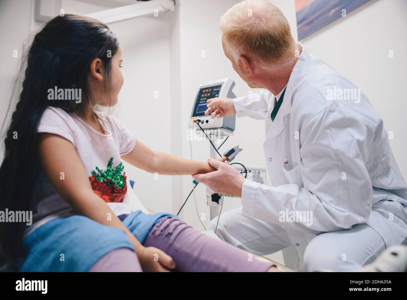 Mature doctor pointing at computer monitor to girl during pulse test in hospital Stock Photo
