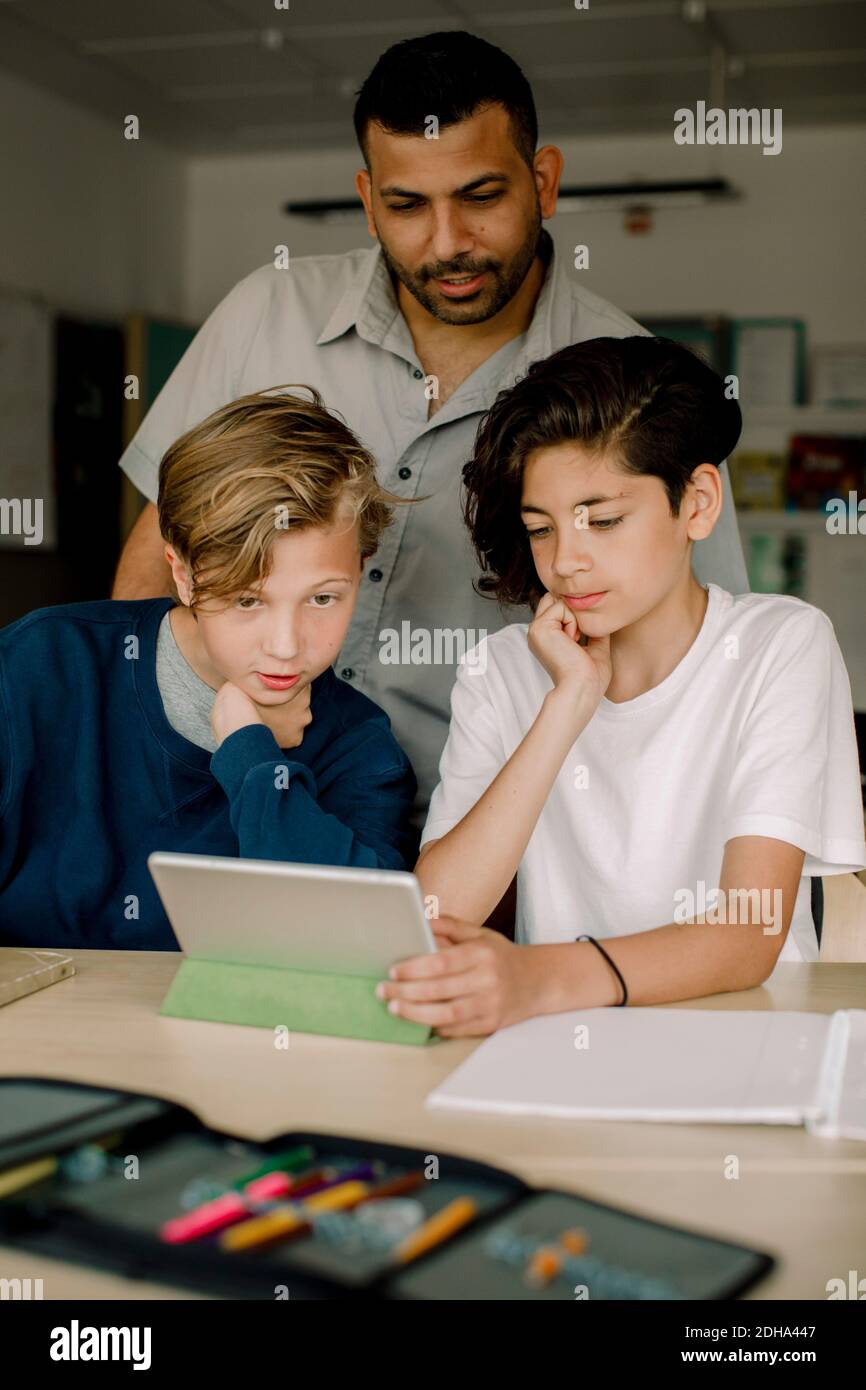 Male tutor with students using digital tablet while sitting in classroom Stock Photo