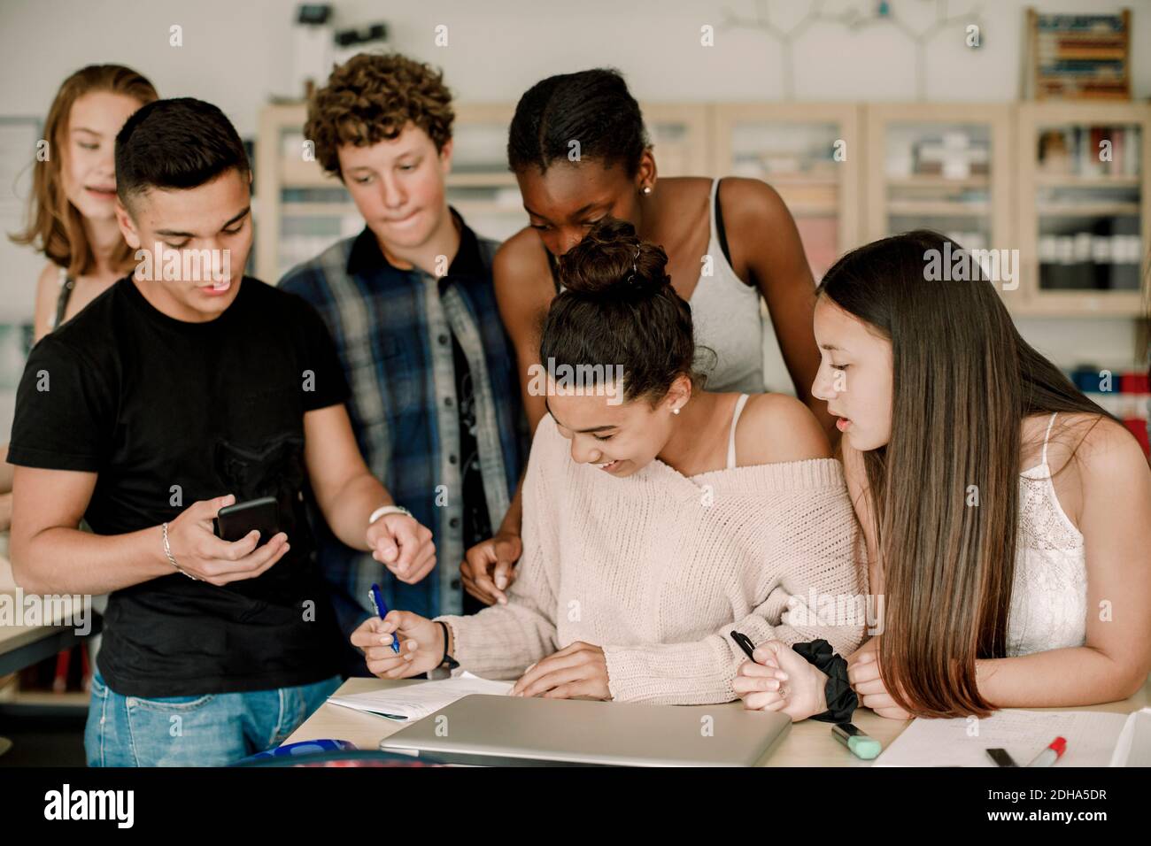 Smiling teenager studying while friends standing by table in classroom Stock Photo