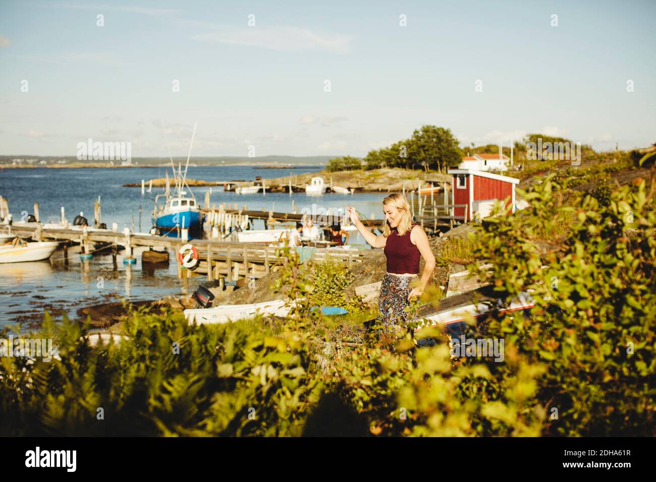 Smiling young woman walking by plants at harbor against sky on sunny day Stock Photo