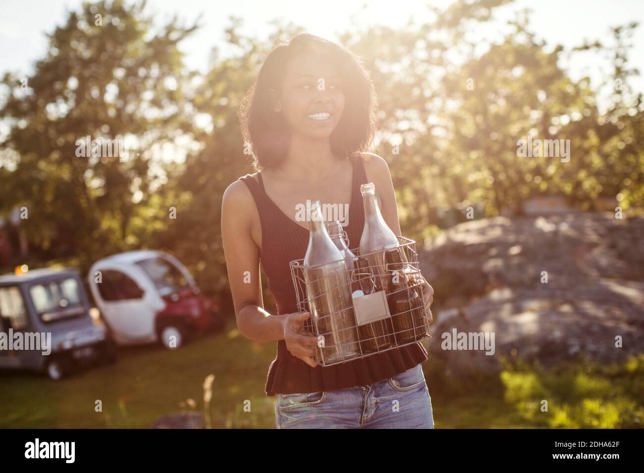 Tilt shot of smiling young woman holding bottles while standing against trees Stock Photo