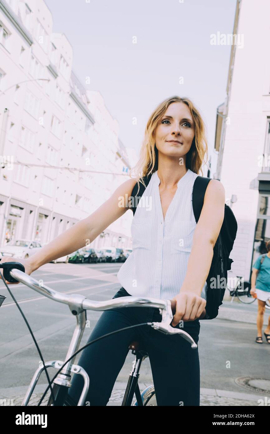 Smiling businesswoman riding bicycle on road in city against sky Stock Photo