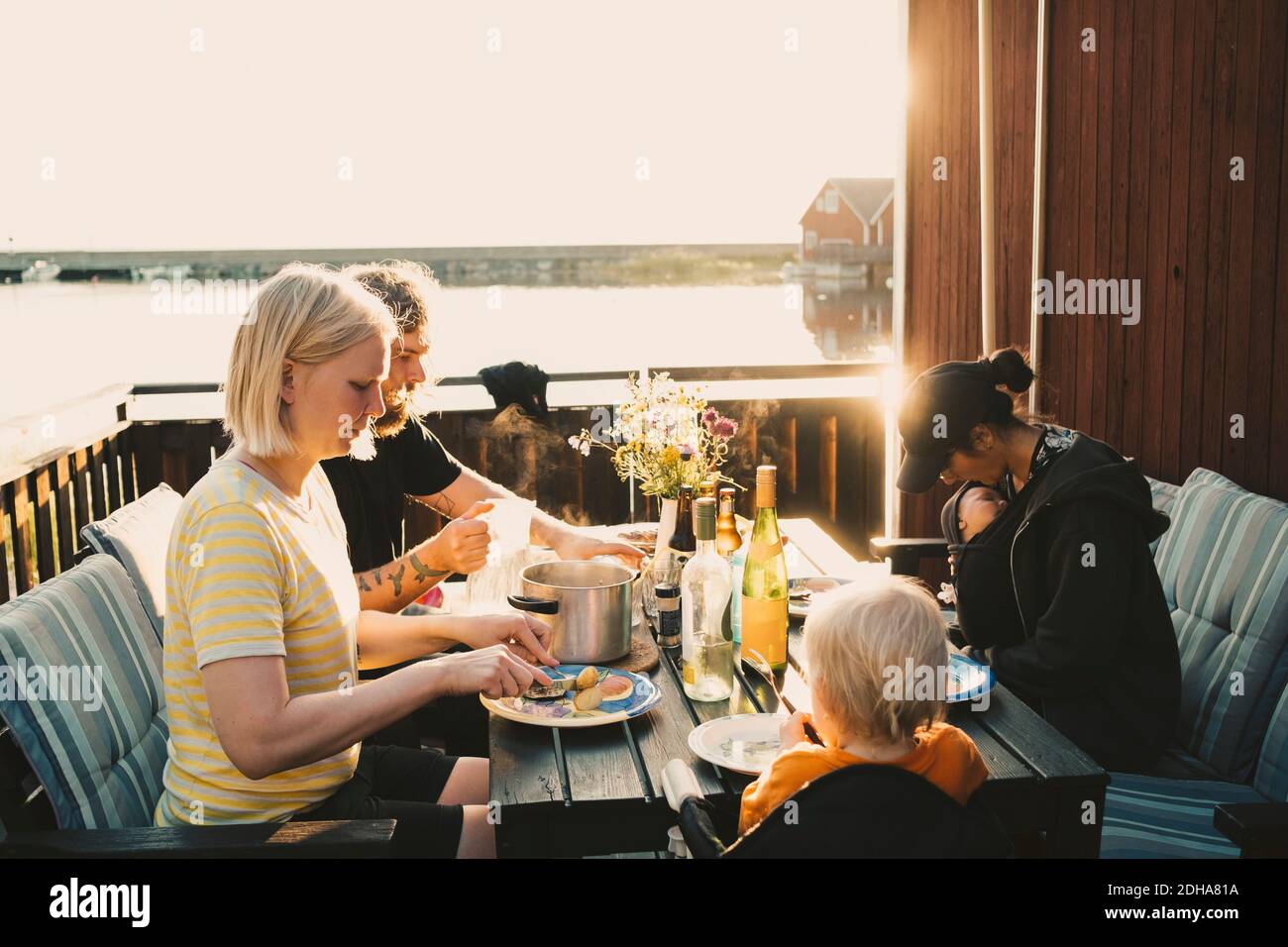 Family and friends having food at table in holiday villa by lake on sunny day Stock Photo