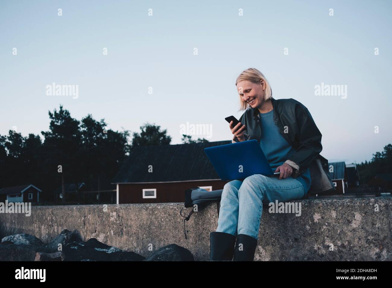 Smiling woman looking at mobile phone and holding laptop while sitting on retaining wall Stock Photo