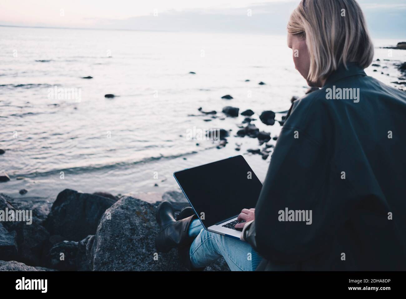 Rear view of woman using laptop while sitting on rock at lakeshore Stock Photo