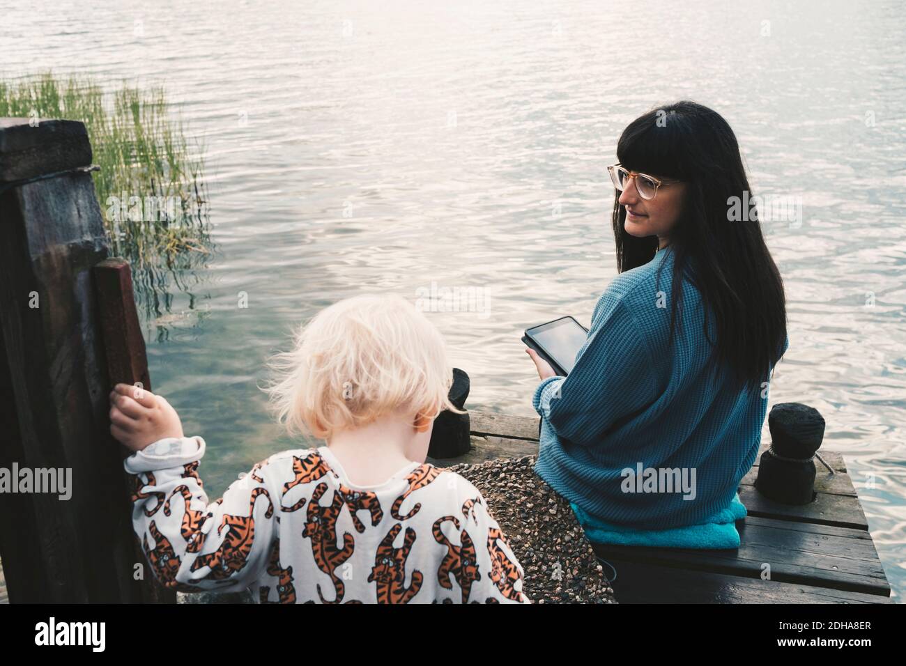 Mother looking at daughter while holding digital tablet and sitting on jetty over lake Stock Photo