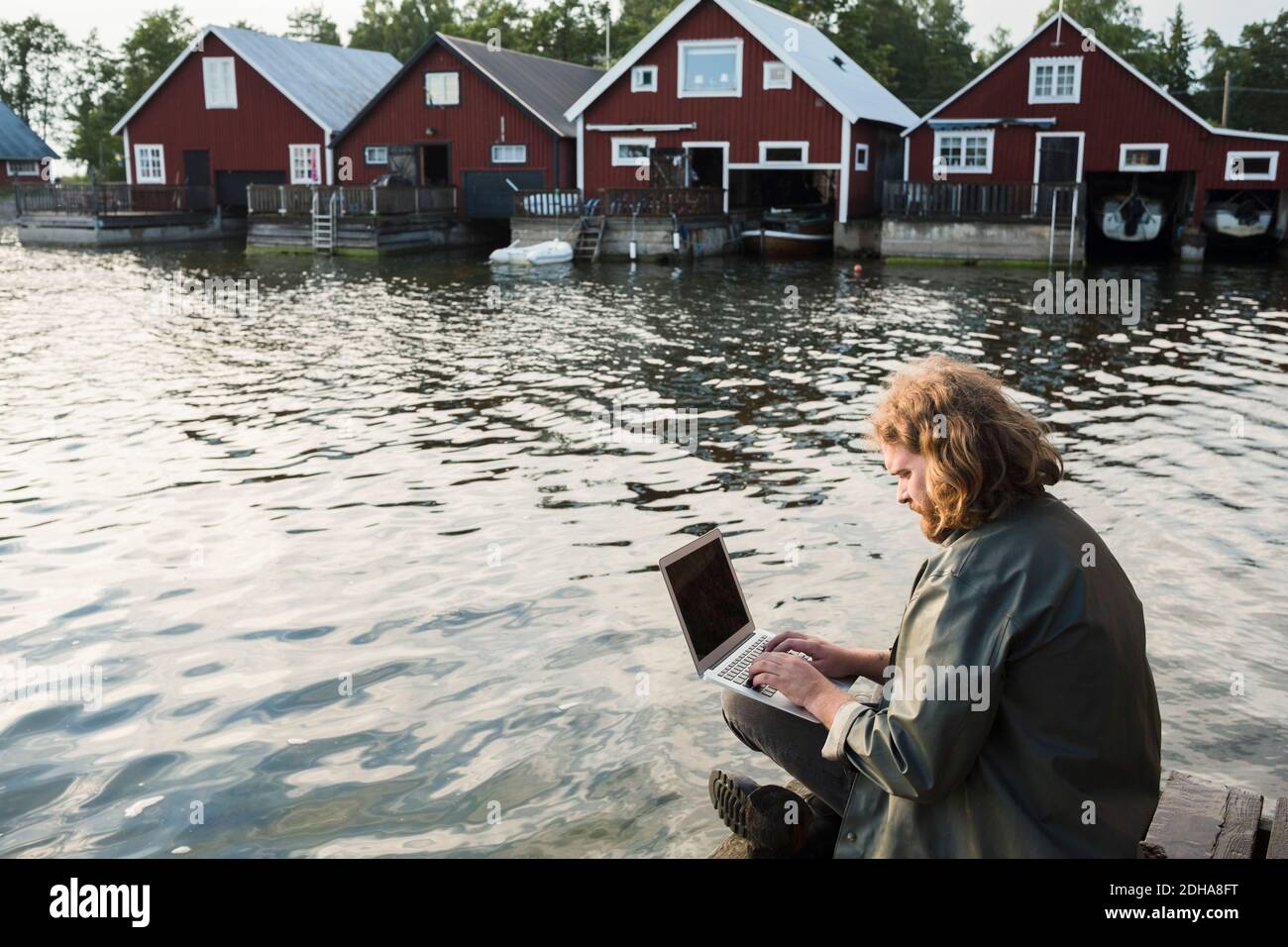Man using laptop while sitting on jetty by lake against holiday villas Stock Photo