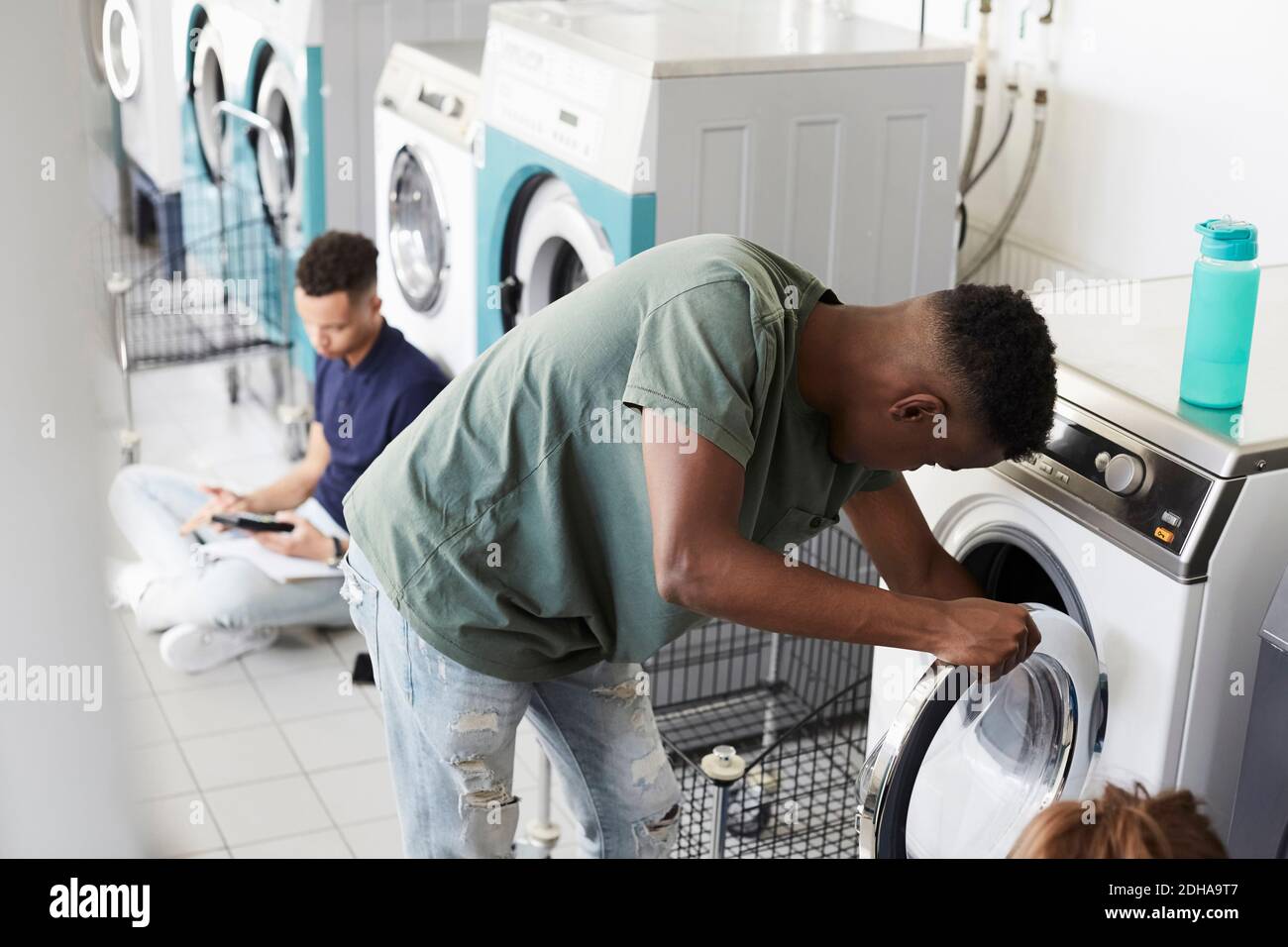 Man using washing machine while university student studying at laundromat Stock Photo