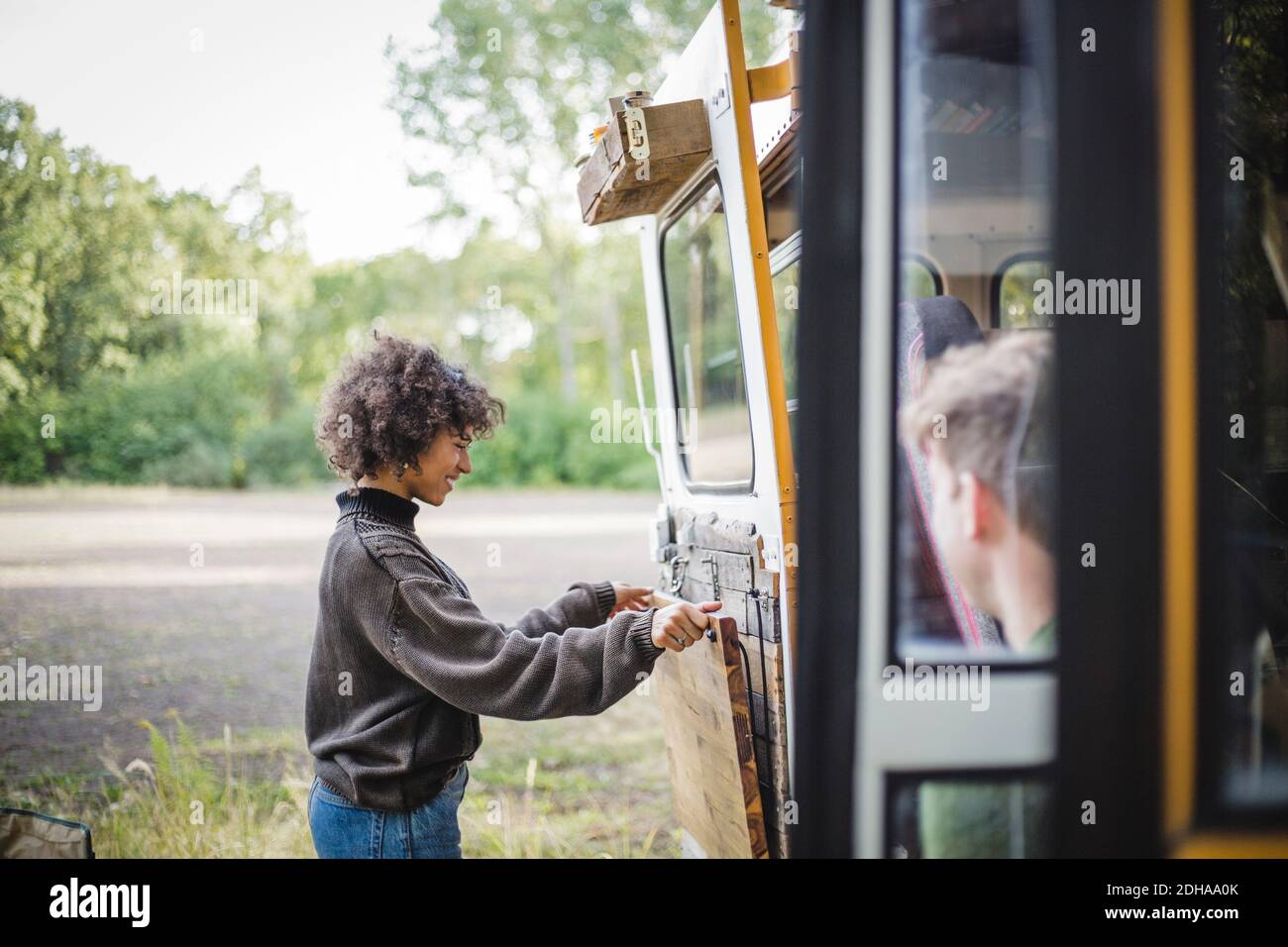 Side view of smiling woman adjusting wood on caravan door during camping in forest Stock Photo