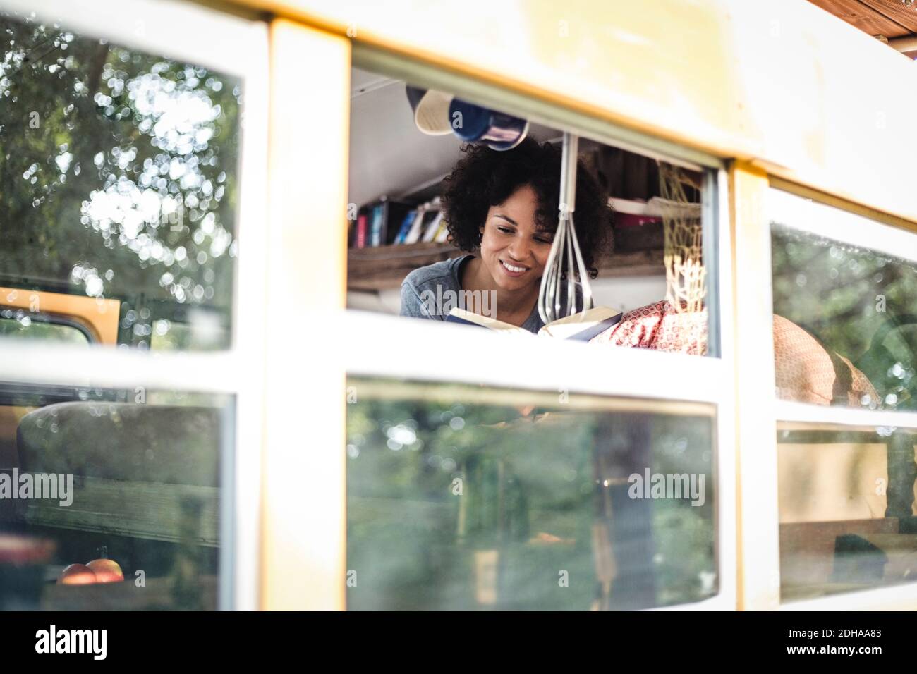 Young Afro woman reading book while lying in motor home seen through window Stock Photo
