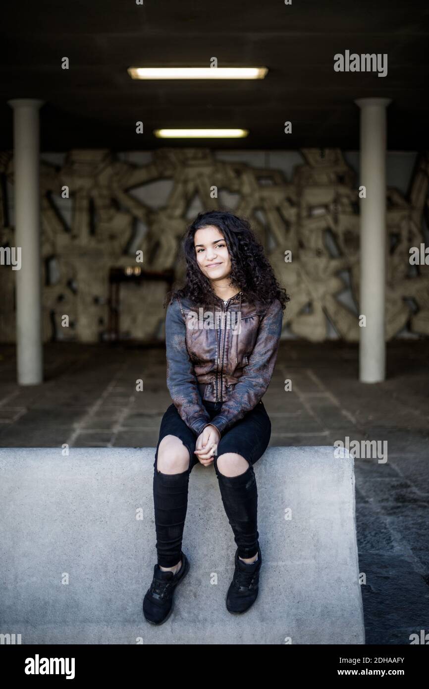 Full length portrait of teenage girl sitting on concrete at parking garage Stock Photo