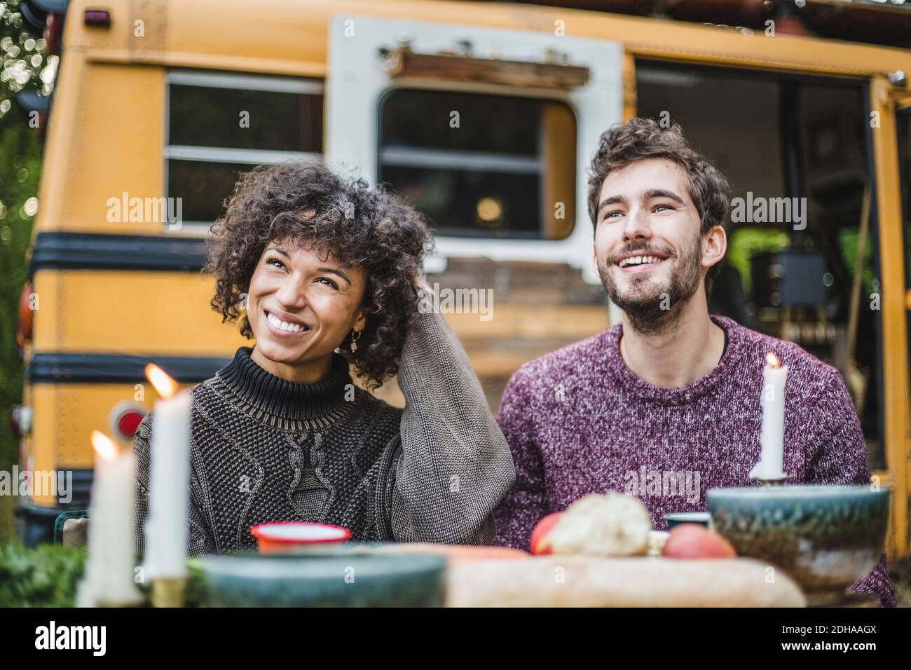 Smiling young couple looking away while sitting at table against caravan during camping Stock Photo
