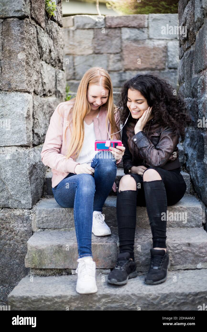 Cheerful female friends sharing smart phone while sitting on steps Stock Photo